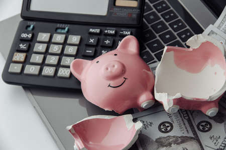 Broken pink piggy bank, calculator and dollar bills on laptop. Finance and bankruptcy conceptの写真素材