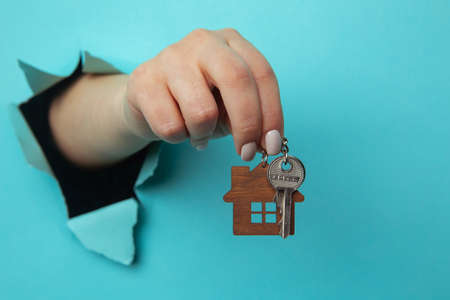 Womans hand with house keys through a hole in blue paper close-up. House sale and rent concept