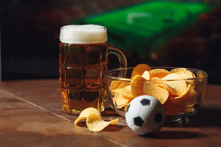 A glass of beer on a wooden table with potato chips. Football on a backgroundの写真素材