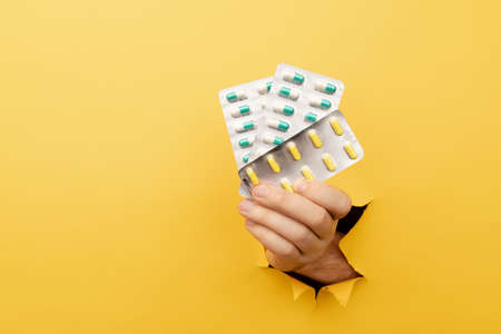 Hand showing a blister of pills out of a hole torn in yellow paper wall. Health care, pharmaceutics...