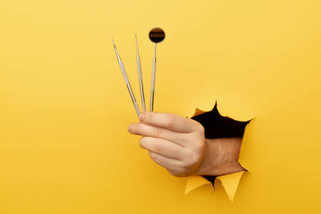 Hand showing a dentist tools out of a hole torn in yellow paper wall. Health care conceptの写真素材