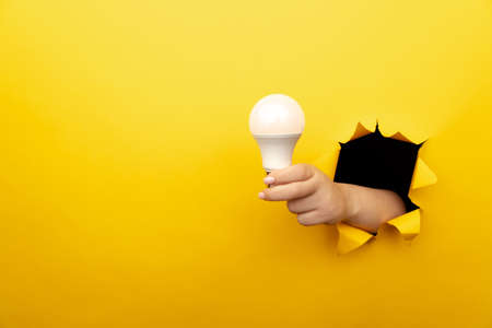 Hand holding an incandescent led light bulb from a torn hole in yellow paperの写真素材