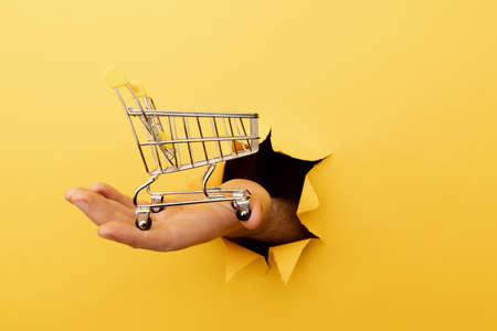 Hand holds through a hole a mini grocery shopping trolley on a yellow paper background. Sales or shopping conceptの写真素材