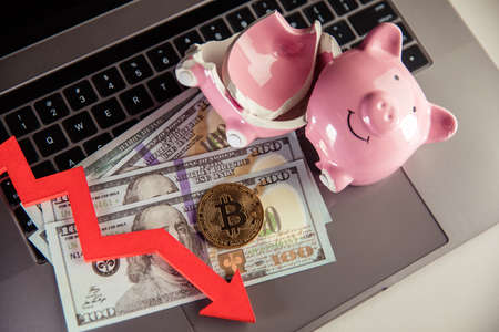Bitcoin, broken piggy bank and arrow down on dollar cash. Cryptocurrency and investment theme. The...