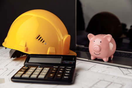 House building costs concept. Safety helmet and piggy bank with blueprint, calculator and laptop on...