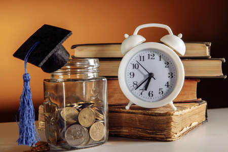 Jar with coins, graduation hat and white alarm clock. Time to save for education conceptの写真素材