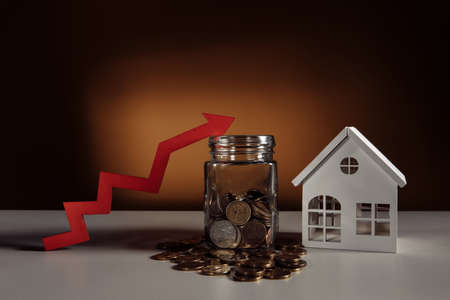 Glass jar filled coins, a red up arrow and house. The concept of the rising price of real estateの写真素材