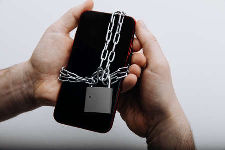 Modern smartphone with chain locked by padlock in mans hand. Information security or smartphone addiction conceptの写真素材