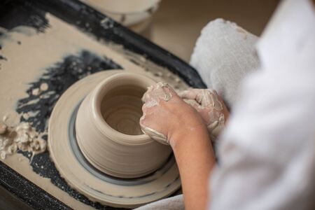 The hands of a professional who creates a pot of clay on a potters wheel. Close-up, top view. Pottery workshop, process of work.の写真素材