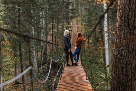 A young albino guy and a red-haired girl stand on in the woods on a suspension bridge and look at...