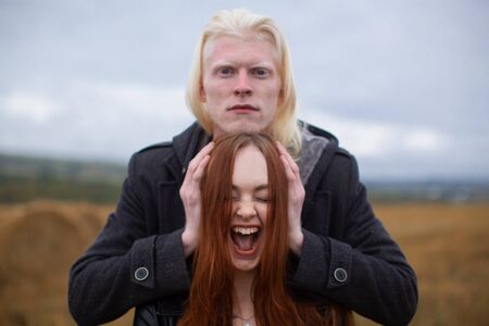 A handsome male albino model with an emotionless face in a black jacket holds his hands on the sides of a red-haired girl who screams against a wheat field. Unusual couple.の写真素材