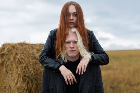 Portrait of a couple. An albino guy with blue eyes in a black jacket and a girl with bright red hair puts her head on his top against the background of a haystack in a field.の写真素材