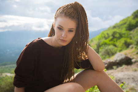 Portrait of a young girl of unconventional appearance with red african braids, provocative makeup in a brown t-shirt against a blue sky and a mountain slope under the scorching rays of the sunの写真素材