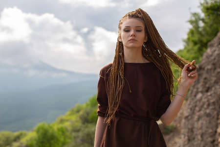 A red-haired girl with cornrows in a brown T-shirt-dress, defiantly looks into the camera holding a strand of hair in her hand and posing against the backdrop of a mountainside and wooded areaの写真素材