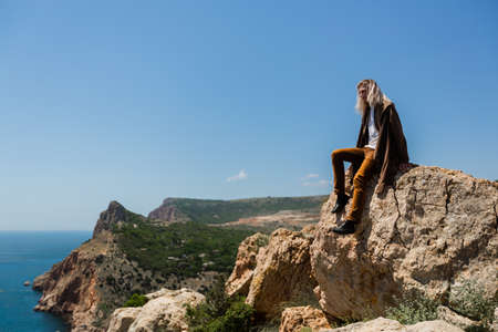 A young man with blond hair up to his shoulders in a brown jacket and orange pants, sits on the edge of a cliff and looks into the distance against the backdrop of a mountain valley and seaの写真素材