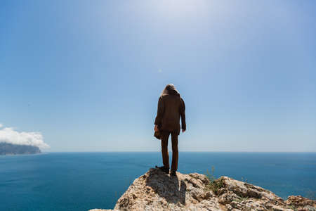 In the foreground there is a man stands on the edge of the cliff with his back to the camera, looking down curiously. A blond man in a brown jacket on the edge of a cliff looks down warilyの写真素材