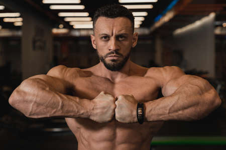 A handsome, handsome bodybuilder with swung back muscles and hands poses looking at the camera and joining his fists at chest level against the backdrop of the gymの写真素材