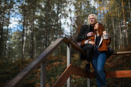 A beautiful young couple sits on railings surrounded by forest. Guy albino with a red-haired girl in love looking at him enjoy a walk in the woodsの写真素材