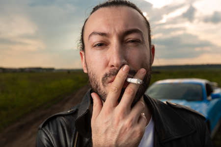 A young brutal man stands on a sandy road smoking a cigarette and looks straight into the cameraの写真素材