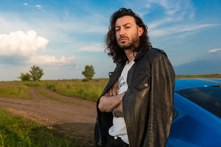 A young brutal man with long hair and bristles stands near a bright blue car on a middle of a rural...