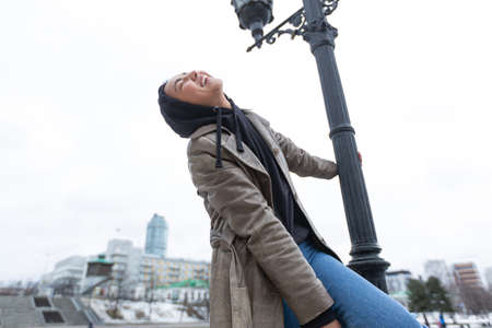 A cheerful girl in a hood and beige coat stands on a black lantern laughing looking up against the...