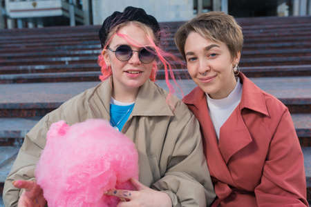 Modern young female hipsters sit on a city street, hold a bright pink cotton wool and smile straight into the camera. High quality photoの写真素材