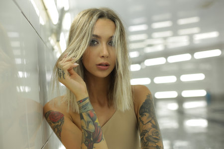 The bright model with blond hair and tattoos on her body stands indoors leaning on the wall and looks directly into the camera. High quality photoの写真素材