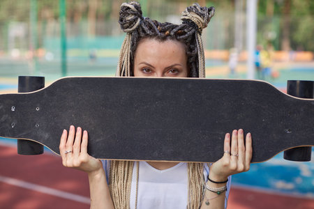 A teenage girl in a white T-shirt and with dreadlocks holds a longboard directly covering her face, staring into the camera. Teenage photo shoot. High quality photoの写真素材