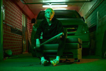 a girl who looks like a guy sits with a bat in her hands near her car in the garage. Shooting in neon green light. High quality photoの写真素材