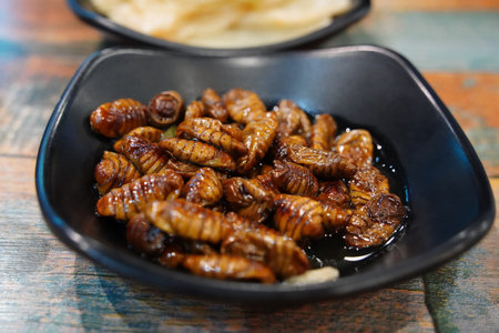 Fried silkworm pupae in a black plate on a wooden table.の写真素材