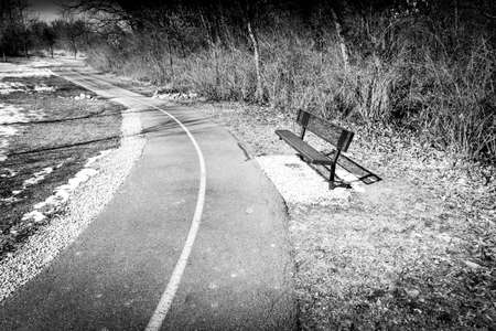 A black and white image of an empty bench near a hiking and biking trailの写真素材