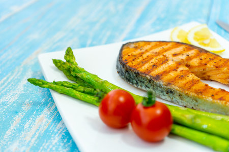 Grilled salmon steak with asparagus and tomato on a plate on blue wooden backgroundの写真素材