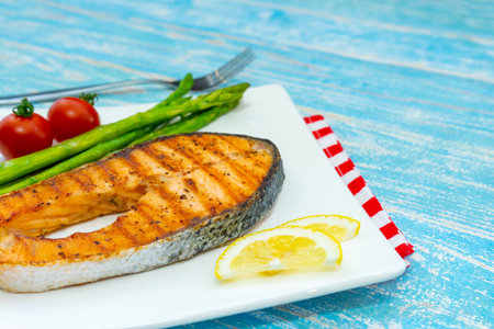 Grilled salmon steak with asparagus and tomato on a plate on blue wooden backgroundの写真素材
