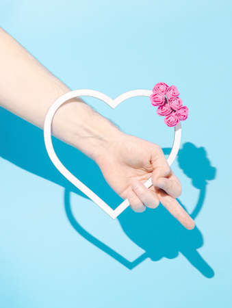 Hand holding white heart frame decorated with pink roses, showing middle finger. Pastel blue background. Minimal creative idea.の写真素材