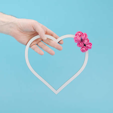 Hand holding white heart frame decorated with pink roses. Pastel blue background. Minimal love concept.の写真素材