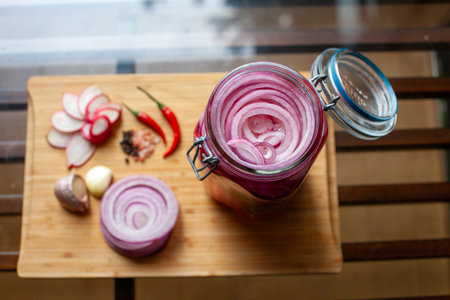 Homemade sweet and spicy pickled red onions sliced in a large glass jar, with the ingredients beside on a wooden cutting board with chili peppers, garlic, radish, rosemary and vinegarの写真素材