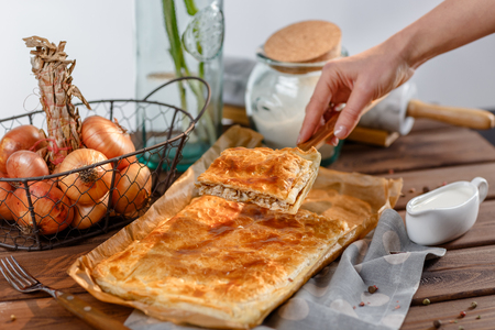 A female hand holds a piece of a square pie on a wooden kitchen spatula. A square piece of meat pie on a rectangular meat pie on a wooden surface against the background of the ingredients from which it was made.の写真素材