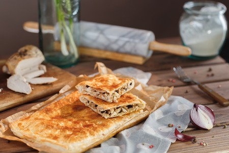 A square piece of meat pie on a rectangular meat pie on a wooden surface against the background of the ingredients from which it was made. Puff pastry cake.
の写真素材