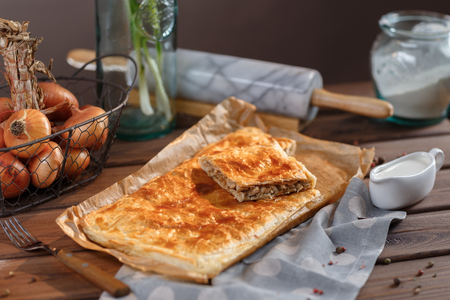 Delicious meat pie on the table with fork bulbs and greens. A square piece of meat pie on a rectangular meat pie on a wooden surface against the background of the ingredients from which it was made.の写真素材
