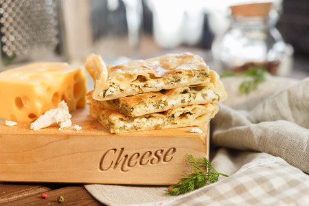 Cheese square cake with cheese and parsley and greens next to a large and juicy piece of yellow cheese Puff pastry cake on a wooden stand central shoot.の写真素材