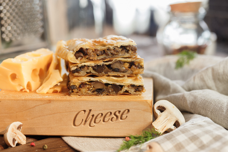 Cheese square cake with mushrooms cheese and parsley and greens next to a large and juicy piece of yellow cheese cut into slices on a wooden stand.の写真素材