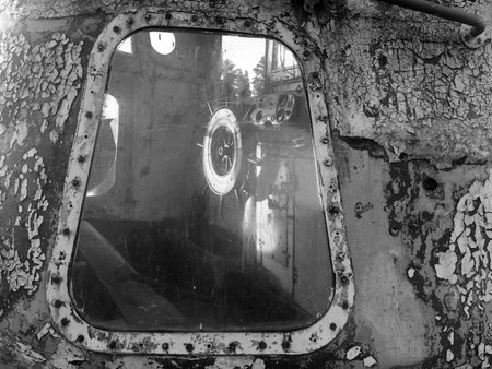 This photo displays a close-up view of a weathered surface with peeling paint, centered around a window. Through the window, there is a reflection showing the interior of an old, abandoned vehicle or machinery, featuring visible dials and controls. The overall ambiance of the image conveys decay and neglect.の写真素材