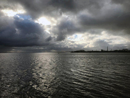 The image depicts a large body of water with ripping waves under an overcast sky. Dark, heavy storm clouds dominate the scene, with patches of light breaking through to illuminate parts of the water. A distant shoreline is visible, featuring industrial structures, including buildings and a tall smokestack. The overall composition captures a dramatic and moody atmosphere, highlighting the contrast between light and shadow.の写真素材
