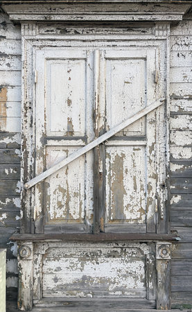 Close-up of an old wooden window with peeling white paint and a diagonal wooden plank nailed across, showing weathered texture and signs of decay.の写真素材