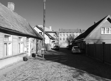 Black and white photo of a narrow cobblestone street with old wooden houses, parked cars, and a distant brick building under a clear sky.の写真素材
