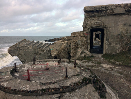 Overcast coastal military ruins: Weathered concrete bunkers and fortifications with arches, battered...