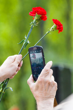 BERLIN - MAY 08, 2015: Victory in Europe Day. Treptower Park. Red carnations and mobile phone. Member of the memorable events photographs at arm's length.のeditorial素材