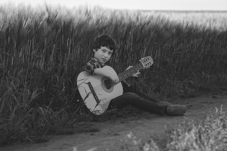 A boy in jeans and a shirt is sitting in the field with a guitar. Black and white. Matte stylization.の写真素材