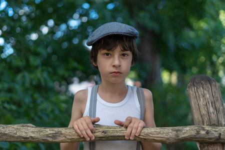 Portrait of a boy dressed in pants with suspenders and a sleeveless shirt near a wooden fence.の写真素材