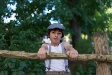 Portrait of a boy dressed in pants with suspenders and a sleeveless shirt near a wooden fence.